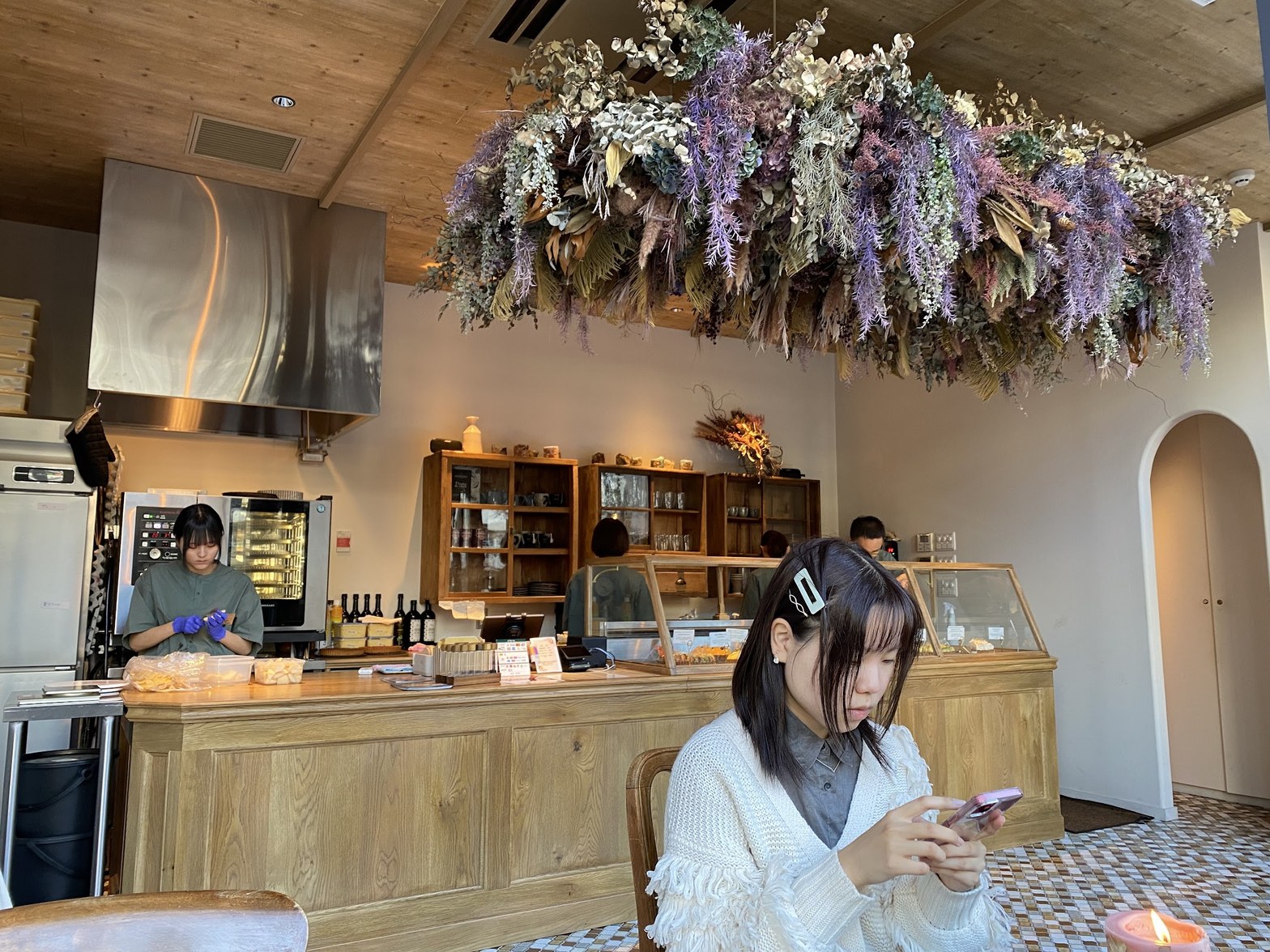 天井の紫と白のドライフラワーを背景に、木製カウンターで作業するスタッフと手前に座る客